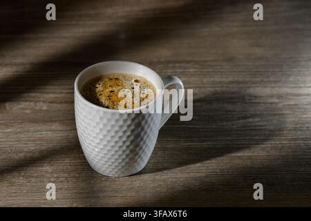 An atmospheric horizontal photograph showcasing a white ceramic mug with a dimpled pattern, filled with freshly brewed black coffee topped with a deli Stock Photo