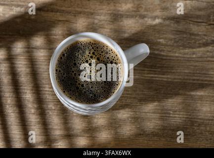 An overhead lifestyle photograph featuring a white ceramic mug with a dimpled pattern, filled with freshly brewed black coffee and a rich layer of bub Stock Photo