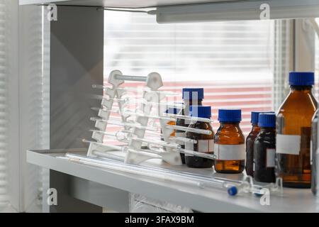 A rack of volumetric glass pipettes and brown reagent bottles with blue caps on a laboratory shelf. Stock Photo