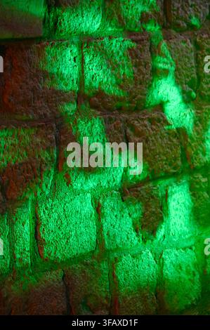 A vertical night photograph capturing a rustic ceramic wall with detailed blocks and grout lines, dramatically illuminated by bright green ambient lighting creating an abstract artistic pattern. Stock Photo