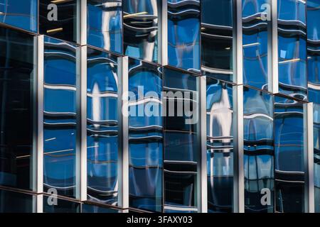 Repeating grid of curved, undulating window panes that fracture the surrounding cityscape into a series of fluid, distorted reflections. Stock Photo