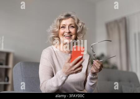 Happy senior woman holding smartphone and eyeglasses home portrait Stock Photo
