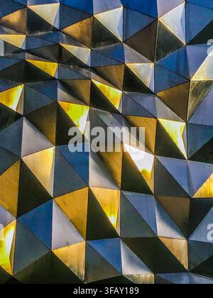 Modern geometric architectural facade with 3D metallic triangular panels reflecting golden light. Stock Photo