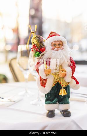 A decorative Santa Claus figurine stands on a table. The Santa has a white beard, red suit, and holds bells and a sack. The background is softly blurr Stock Photo