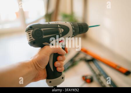 Hand holding an electric drill with bit poised to drill into a wall, tv bracket and tools blurred in background diy home renovation and mounting in progress Stock Photo