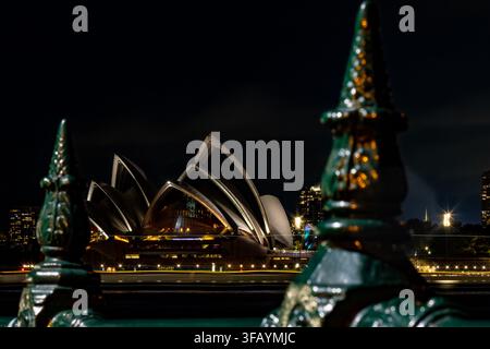 Night photograph of the Sydney Opera House with its illuminated sails, framed between two out‑of‑focus green decorative fence finials. Stock Photo