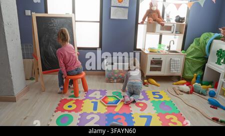 Girl draws on chalkboard while another child kneels on a number puzzle mat and stacks toys in an indoor playroom building; playful learning. Stock Photo