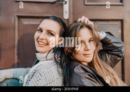 Women friendship portrait of two female friends sitting back to back smiling outdoors in warm sunset light Stock Photo