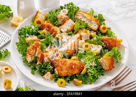 Sliced breaded pork cutlet salad with green olives, grated parmesan cheese and fresh lettuce leaves served on a plate over white marble table with for Stock Photo