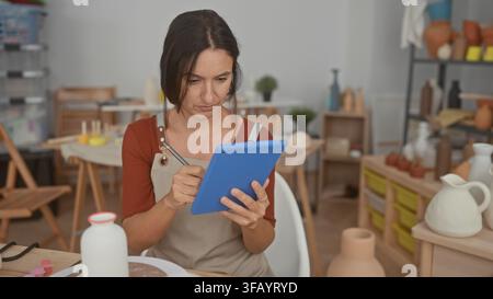 Woman holds blue tablet and taps with stylus in pottery studio, hands visible and focused on screen while surrounded by shelves of ceramic vases; crea Stock Photo