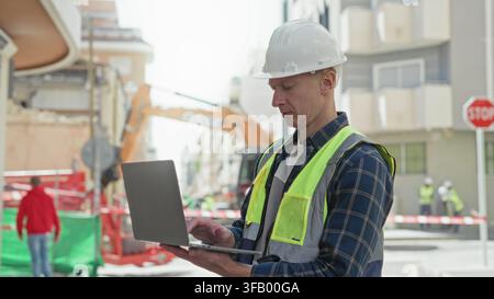Man builder in hardhat and high visibility vest holding laptop and inspecting a street construction site; safety planning focus. Stock Photo