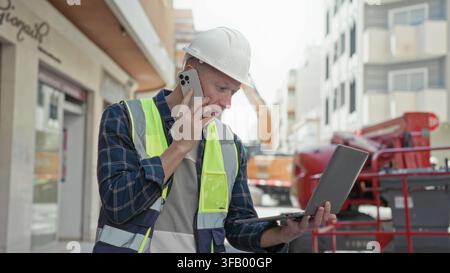 Man builder in hardhat and hi vis vest talks on phone while holding laptop on an urban street with excavator behind him; focus duty. Stock Photo