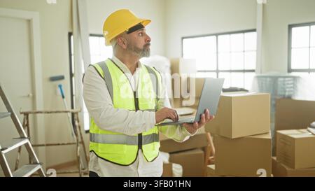 Man typing on laptop with hands visible in a building renovation room, wearing hardhat and safety vest beside ladder and stacked cardboard boxes; prod Stock Photo