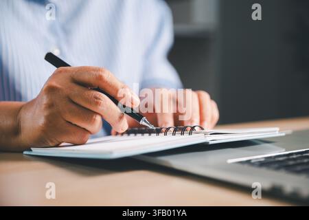 Close-up of a person writing notes in a spiral notebook placed on a laptop, representing online learning, academic research, remote study, productivit Stock Photo