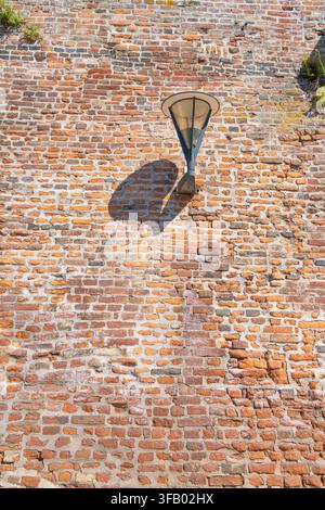 A modern lantern is mounted on a textured, weathered brick wall. Sunlight casts a distinct shadow of the lantern onto the wall. Stock Photo