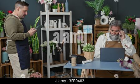 Two men florist workers in aprons, one holds a tablet while the other talks on a smartphone and uses a laptop at a shop building; teamwork focus. Stock Photo