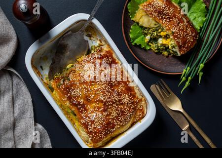 Top down view of freshly baked savory pastry pie filled with greens and boiled eggs in ceramic baking dish with cut piece on plate, close up Stock Photo