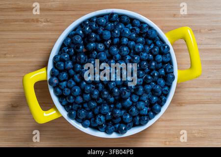 Fresh wild blueberries in white bowl with yellow handles. Top view of white ceramic bowl filled with ripe wild blueberries or bilberries, placed on li Stock Photo