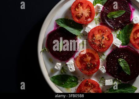 Top view of gourmet salad featuring sliced beetroot, red tomatoes, feta cheese cubes, and mint leaves with olive oil and spices on white plate, isolat Stock Photo