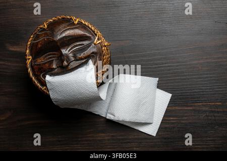 Funny wooden mask toilet paper holder on dark background, close up. Unique hand carved wooden face tissue dispenser with white toilet paper roll on da Stock Photo
