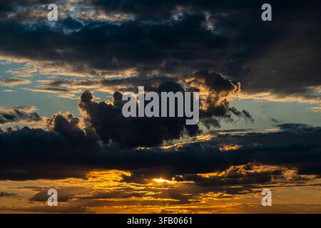 Golden sunset behind dramatic dark clouds. Sunset sky with bright orange light breaking through heavy dark storm clouds. Perfect for weather forecasti Stock Photo