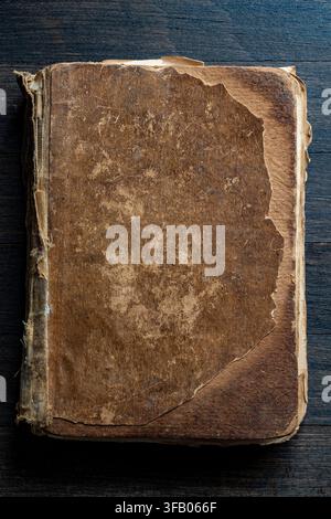Vintage worn book cover on dark wood, close up. Top view of an antique book with heavily damaged, weathered brown leather cover lying on dark wooden b Stock Photo