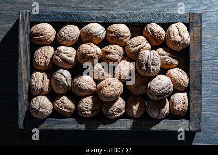 Whole walnuts in dark wooden box. Top view of raw whole walnuts in shells neatly arranged in rustic dark wooden tray crate on table, dramatic side lig Stock Photo