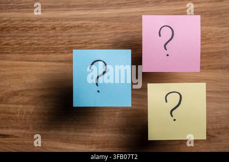 Top view of three colorful paper sticky notes with handwritten black question marks on textured wooden office desk background, close up. Choice decisi Stock Photo