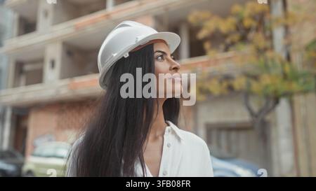 Woman architect in hardhat with hand on chin looking up at building, profile pose and slight smile; thoughtful confidence. Stock Photo