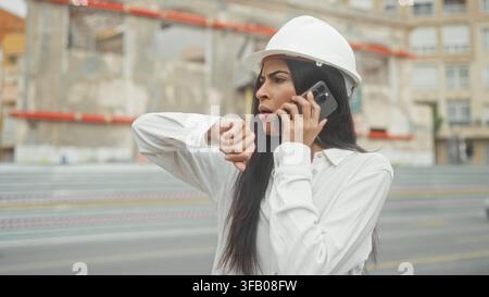 Woman architect in hardhat on building site talks on smartphone to ear while checking wristwatch and studying plans in hand; deadline urgency. Stock Photo