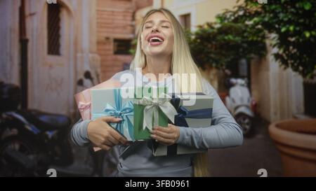 Young blonde woman holding several wrapped gifts with hands to chest, eyes closed and smiling on a narrow street with scooters and a potted tree; joy Stock Photo