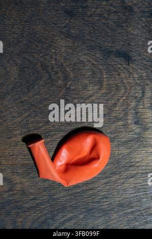 Deflated orange balloon on dark wooden background, close up. Single flat deflated orange rubber balloon lying on textured dark wood surface with copy Stock Photo