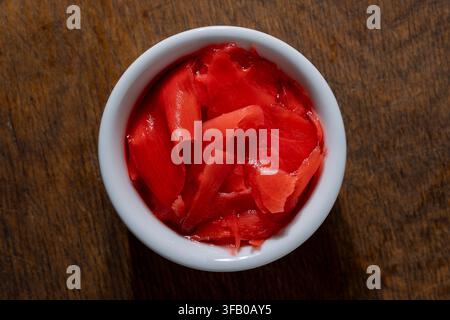 Pickled pink ginger slices in white ceramic bowl, close up. Top view of small white round bowl filled with bright pink pickled ginger gari placed on r Stock Photo