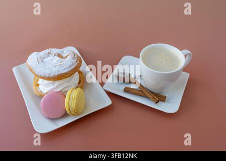 Elegant still life with white cup of cappuccino coffee, sweet colorful macarons, crispy meringue cookies and cinnamon sticks on clean table Stock Photo
