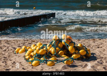 A big heap of bright yellow commercial fishing floats tangled with blue ropes lies on fine sand. Foamy waves of the Baltic Sea crash against a long wo Stock Photo