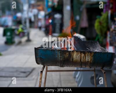 Rustic charcoal brazier with burning fire and glowing embers on a city sidewalk in Southeast Asia, busy street scene blurred behind. Stock Photo