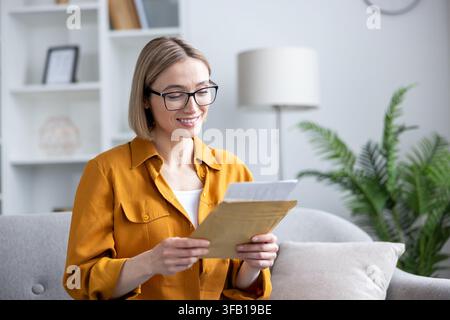 Smiling woman wearing glasses opening an envelope, reading a letter with good news, feeling happy and positive while sitting on a sofa in a bright modern living room Stock Photo