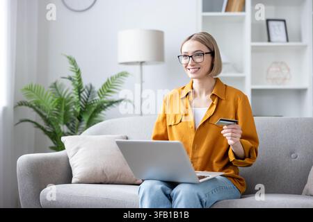 Young woman wearing glasses and a yellow shirt sitting comfortably on a gray sofa, smiling happily while using her laptop and holding a credit card for convenient online shopping at home Stock Photo