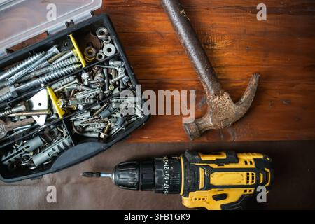 Top view of a yellow cordless drill, worn claw hammer, and open case filled with screws and hardware on a wooden workbench. Stock Photo