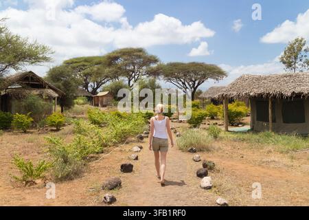 Rear view of a female tourist walking down a stone-lined dirt pathway toward thatched-roof eco huts and acacia trees in a safari lodge setting in Africa Stock Photo