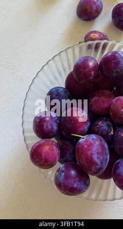 Ripe purple plums arranged in a clear glass bowl, creating a simple fresh fruit still life on a light surface. Stock Photo
