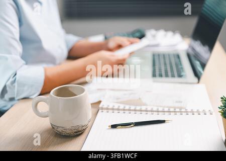 Coffee mug, smartphone, notebook, and laptop on an office desk while a business professional reviews documents, representing productivity, planning, c Stock Photo