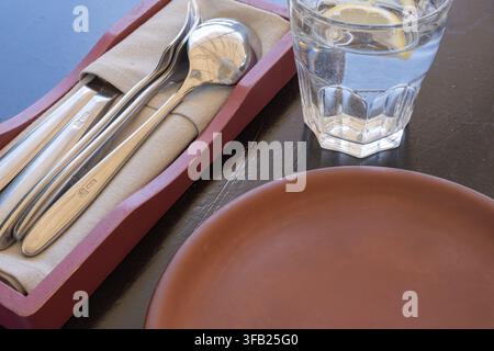 Restaurant table prepared with an empty ceramic plate, cutlery on a napkin and a glass of lemon water Stock Photo