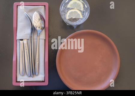 Restaurant table prepared with an empty ceramic plate, cutlery on a napkin and a glass of lemon water Stock Photo