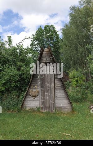 Weathered wooden garden shed with closed plank door, framed by tall green trees, lawn, shrubs, and cloudy sky in quiet rural yard  Stock Photo