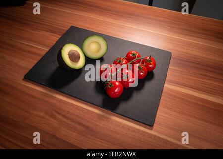 High-angle food still life featuring avocado halves and a cluster of fresh cherry tomatoes on the vine against a black cutting board. Stock Photo