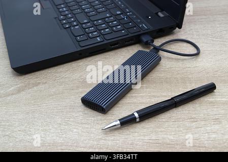 High-angle view of a grey ribbed external NVMe solid state drive plugged into a laptop using a USB cable, featuring a sleek black ink pen resting on a Stock Photo