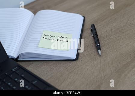 Close-up of a yellow adhesive sticky note with login credentials and a password written on it, attached to a black notebook on a wooden office desk. L Stock Photo