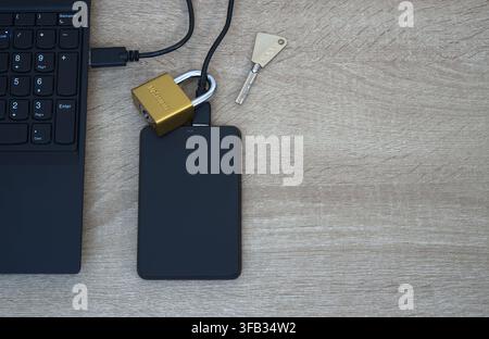 Close-up view of a small brass padlock physically secured around the USB cable of a black external hard drive. Laptop keyboard visible on the side. Co Stock Photo