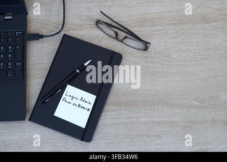 Close-up of a yellow adhesive sticky note with login credentials and a password written on it, attached to a black notebook on a wooden office desk. L Stock Photo